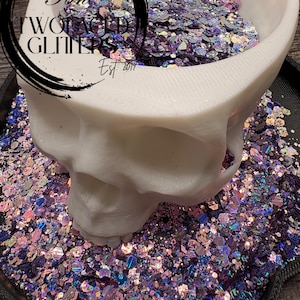 May include: A white skull-shaped container filled with iridescent glitter in shades of purple, pink, and blue. The glitter is scattered around the base of the container. The container has the text "Two Faced Glitters Est. 2017" in a black circle.