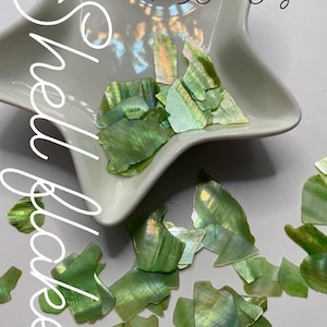 May include: Green shell flakes in a white, star-shaped dish. The iridescent flakes have a shimmering, pearlescent quality. The word "Shell" is written in a cursive font on the left side of the image.