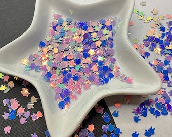 May include: A white star-shaped dish filled with iridescent pastel blue, pink, and purple bunny-shaped glitter. The glitter is scattered around the dish on a white surface.