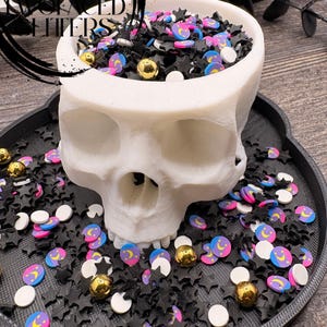 May include: A white skull-shaped container filled with black star-shaped sprinkles, gold spheres, and colorful moon and star-shaped sprinkles. The container sits on a black tray, surrounded by more sprinkles. The theme is Halloween.