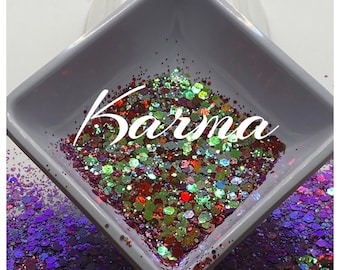 May include: A close-up of a small white square dish filled with colorful iridescent glitter. The glitter is a mix of red, green, gold, and blue. The word "Karma" is written in white cursive letters on top of the glitter.