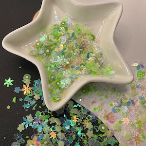 May include: A white star-shaped dish filled with iridescent green, blue, and white glitter. The glitter is in the shape of stars, flowers, and other abstract shapes. The dish is surrounded by more glitter on a white surface.