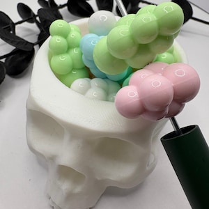 May include: A white skull-shaped container filled with colorful, bubble-like objects in shades of green, blue, pink, and white. Two dark green rods are inserted into the container, adding a contrasting element to the design. The overall theme is a unique, decorative item.