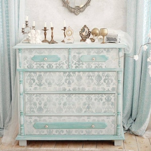 May include: A light blue dresser with four drawers and decorative silver and white patterned paper. The dresser has ornate wooden details and is decorated with candlesticks and other decorative items.