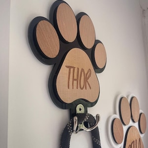 Puede incluir: Gancho de pared de madera con forma de huella de pata negra con el nombre "THOR" grabado. Una correa negra cuelga del gancho.