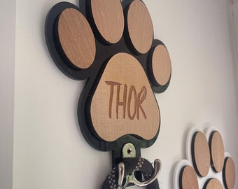 May include: Black paw print shaped wooden wall hook with the name "THOR" engraved on it. A black leash is hanging on the hook.