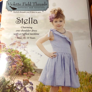 May include: A girl wearing a light blue one-shoulder dress with a ruffled neckline. The dress is made for sizes 2T-10 years. The dress is called "Stella" and is a charming one-shoulder dress with a ruffled neckline. The image is from Violette Field Threads, a company that sells patterns and tutorials for stylish threads.