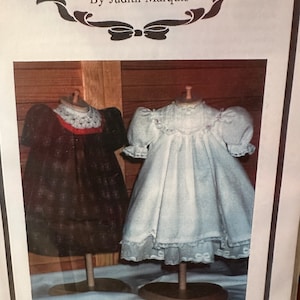 May include: Two doll dresses on display stands. The dress on the left is black and white plaid with a red trim and a white crocheted collar. The dress on the right is white with a lace trim and a white crocheted collar. The text "The Grace Knott Doll Clothes Collection By Judith Marquis" is at the top of the image. The text "Belle" is at the bottom of the image.