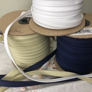 May include: Three spools of fabric trim, one white, one beige, and one navy blue. The spools are on a wooden surface.