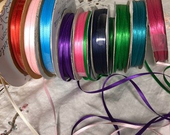 May include: A collection of spools of satin ribbon in various colors, including white, pink, light blue, purple, red, green, teal, and navy blue. The spools are arranged in a circle, with the ribbon ends trailing out.