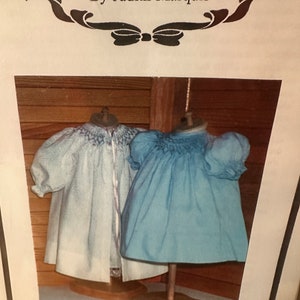 May include: Two doll dresses on display stands. The dress on the left is white with blue embroidery and a blue ribbon. The dress on the right is blue. The text "The Peasant Dress" is visible below the dresses.