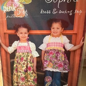 May include: Two girls wearing floral print dresses and denim pants. The girls are standing in front of a window with a chalkboard sign that reads "the Sophia dress & swing top pink fizz patterns".