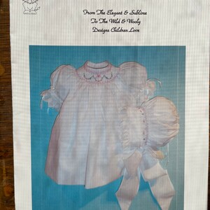 May include: A white baby dress with a pink embroidered design and a matching white bonnet. The dress has puffed sleeves and a gathered skirt. The bonnet has a ruffle trim. The text "295 Angelica" is printed below the image.