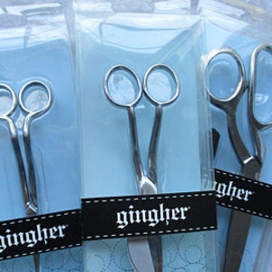 May include: Three pairs of silver scissors in different sizes, all packaged in clear plastic with blue backing cards. The scissors have the word "gingher" printed on the cards.