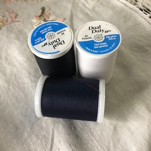 May include: Three spools of Dual Duty XP thread. The spools are white, black, and navy blue. Each spool has the text "Dual Duty XP" and "All Purpose" printed on it. The white spool also has the text "Tout usage" and "Uso general" printed on it.