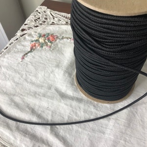 May include: A spool of black elastic cord, wound around a cardboard spool with a paper label. The cord is lying on a white lace tablecloth.