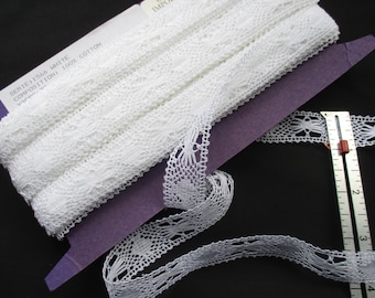 May include: White cotton lace trim with a delicate floral pattern. The label reads "SERIE 1.566 WHITE COMPOSITION 100% COTTON YARDS -"