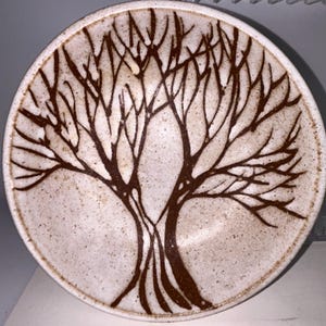 May include: A round ceramic bowl with a speckled off-white glaze featuring a dark brown hand-painted tree design. The tree has a thick trunk that branches out into numerous thin, bare limbs covering the interior surface of the bowl.