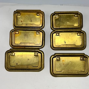 May include: Six vintage brass drawer pulls with a rectangular design. Each pull has a recessed handle and a slightly aged appearance. The pulls are arranged in two rows of three, showcasing their classic hardware style.