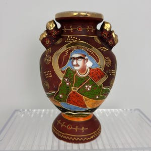 May include: A dark maroon ceramic vase featuring a hand-painted figure in a green and red patterned robe with a blue head covering. The vase has gold-painted accents, two decorative side handles, and a circular base with gold line detailing.