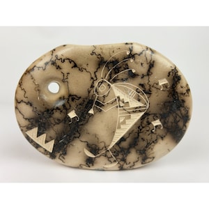 May include: A light brown oval-shaped stone with a black and white design of a stylized figure with a headdress. The design is created using a technique called 'fired-in' or 'smoke-fired' where the design is etched into the stone and then fired, leaving a black outline.