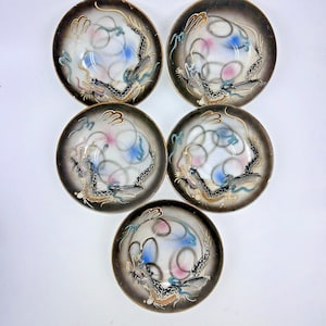 May include: Five circular ceramic saucers featuring a central dragon motif in black, gold, and blue. Each saucer has a white center with soft pink and blue airbrushed accents, surrounded by a dark brown gradient rim and gold-toned edge detailing.