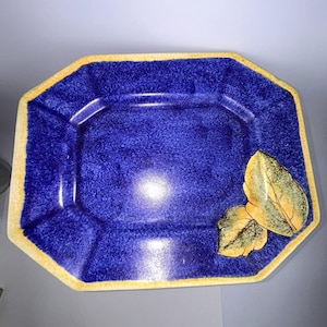 May include: A blue ceramic serving platter with a yellow rim and leaf accents. The octagonal platter has a textured surface and is decorated with two yellow and green leaves on the right side. A decorative piece for serving food.