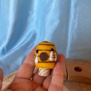 Könnte beinhalten: Eine nadelgefilzte gelb-braune Bienenfigur mit schwarzen Augen und weißen Flügelakzenten, befestigt auf einem kleinen zylindrischen Holzsockel. Die Biene hat einen runden Körper mit horizontalen braunen Streifen und eine flauschige Oberfläche.