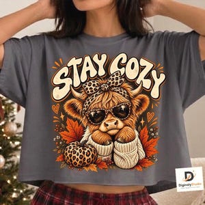 Peut inclure: Un t-shirt gris anthracite avec un motif de vache Highland portant des lunettes de soleil, un nœud à imprimé léopard et un pull crème. Le design inclut le texte STAY COZY, une citrouille à motif léopard et des feuilles d'automne orange.