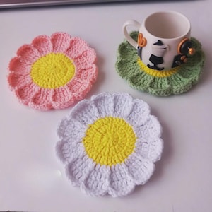 Peut inclure: Trois dessous de verre faits à la main en crochet en forme de fleurs. L'un est rose, l'autre blanc et le dernier vert. Chaque dessous de verre a un centre jaune. Une petite tasse à café blanche avec un motif décoratif repose sur le dessous de verre vert.