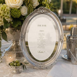 Op de afbeelding: Een ovaal zilverkleurig frame met een gedrukt menu getiteld THE BAR, met een lijst van diverse cocktails zoals Aperol Spritz, Negroni en Espresso Martini, onderaan afgesloten met het woord Salute, geplaatst op een tafel met bloemendecoratie.
