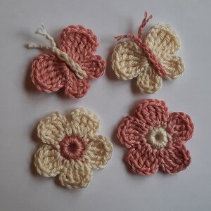 Puede incluir: Cuatro adornos de ganchillo: dos mariposas rosas y dos flores con centros rosas y pétalos crema.