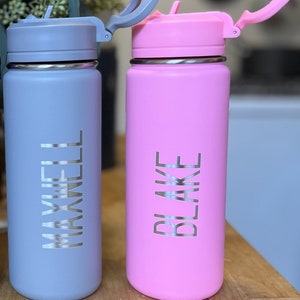 Könnte beinhalten: Zwei Edelstahl-Wasserflaschen mit personalisierten Namen. Die Flasche links ist grau mit dem Namen "MAXWELL" und die Flasche rechts ist rosa mit dem Namen "BLAKE".