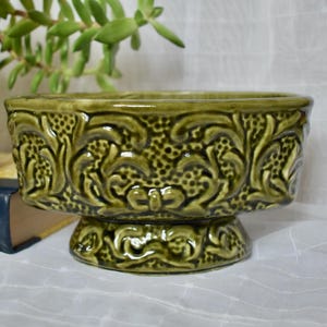 Peut inclure: Un bol en céramique vert olive de forme ronde, orné d'un motif en relief texturé sur toute sa surface extérieure. Le bol repose sur une base en piédestal qui reprend le même design complexe et gaufré que celui du corps principal.