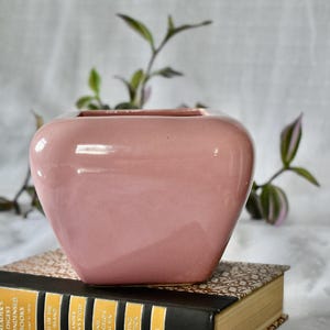 May include: A square-shaped ceramic planter with a smooth, glossy, dusty pink finish. The planter features rounded corners and a wide, open top, presenting a clean, solid-colored surface that reflects light across its curved, bulbous exterior walls.