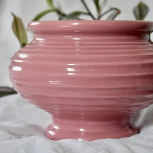 Peut inclure: Un cache-pot en céramique rose poudré avec une finition brillante et des détails nervurés horizontaux. Le récipient présente un corps large et arrondi qui se rétrécit vers une base circulaire, avec une plante visible au sommet.