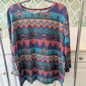 May include: A long-sleeved top with a colorful geometric pattern. The design features zigzags, diamonds, and other shapes in shades of teal, pink, purple, and gold. The top is hanging on a gray hanger.