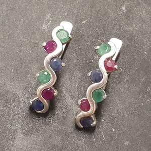 May include: A pair of silver hoop earrings with a wavy design. Each earring features five gemstones: two green, two red, and one blue.