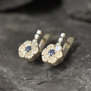 May include: A pair of silver earrings with a flower design. Each earring features a cluster of white moonstone stones surrounding a small blue sapphire.