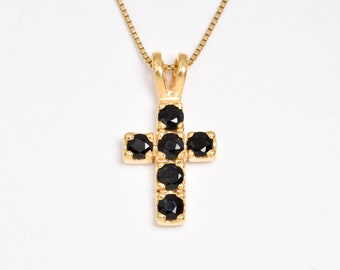 May include: A gold-colored cross pendant necklace with black gemstones. The cross is made of gold-toned metal and features seven round, black gemstones. The necklace has a delicate gold chain and is set against a white background.