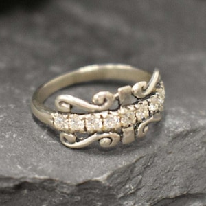 Puede incluir: Un anillo de plata con un diseño delicado y ornamentado que presenta una fila de piedras de circonita cúbica transparentes.