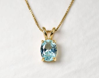 Puede incluir: Un collar de oro con un colgante de piedra preciosa azul claro de corte ovalado, sujeto por un engaste de cuatro garras. El colgante cuelga de una delicada cadena de oro con eslabones cuadrados y presenta un acabado de metal pulido.