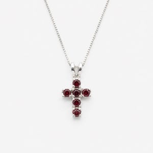 Puede incluir: Un collar de tono plateado con un colgante en forma de cruz engastado con seis piedras preciosas redondas de color rojo intenso. El colgante cuelga de una cadena fina y delicada de tono plateado. El diseño de la cruz es simétrico, con una piedra en la parte superior, una a cada lado y tres piedras formando la base vertical.