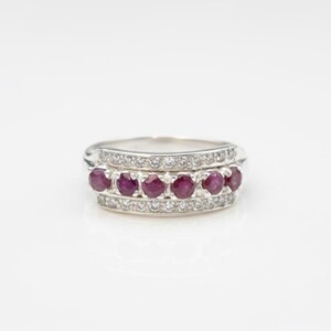 May include: A silver ring featuring a row of six round, deep red rubies set between two rows of small, clear gemstones. The ring has a classic design, with a polished finish, and is set against a white background.