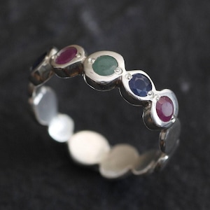 May include: A silver band ring with a row of colorful gemstones. The ring features a green, blue, red, and blue gemstone.