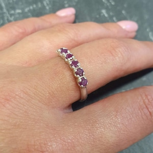 Pode incluir: Um anel de prata com uma fileira de sete pequenas pedras de rubi vermelho escuro engastadas em uma banda curva.