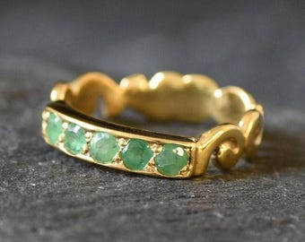 May include: A gold band ring with a row of emerald green gemstones set in a decorative, swirling design.