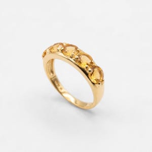 May include: A gold ring featuring five oval-cut, yellow gemstones. The ring has a polished gold band and the gemstones are set in a row across the top. The ring is displayed on a white background.