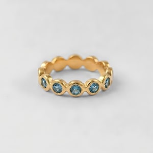 Peut inclure: Une bague dorée ornée d'une série de pierres précieuses rondes bleu clair. La bague présente un design continu, chaque pierre étant sertie dans un sertissage à griffes. La bague est présentée sur fond blanc, mettant en valeur sa couleur et son design. Diamètre d'environ 1,8 cm.