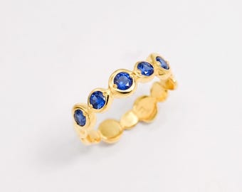 May include: A gold band ring featuring a series of five round, vibrant blue sapphire gemstones set in individual circular bezels. The metal has a polished finish, and the stones are arranged in a row along the top half of the circular band.